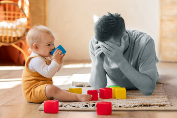 Pai cansado mentalmente, aparentemente com burnout, deitado no chão com as mãos no rosto em frente a seu bebê que brinca sentado.